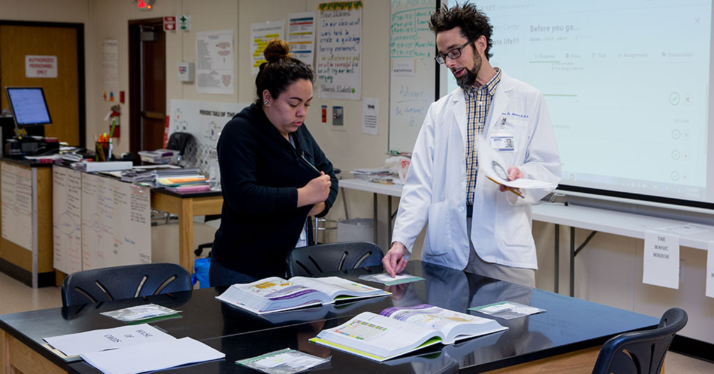 A high school student and science teacher look at a text book together with a smart board in the background of the lab.