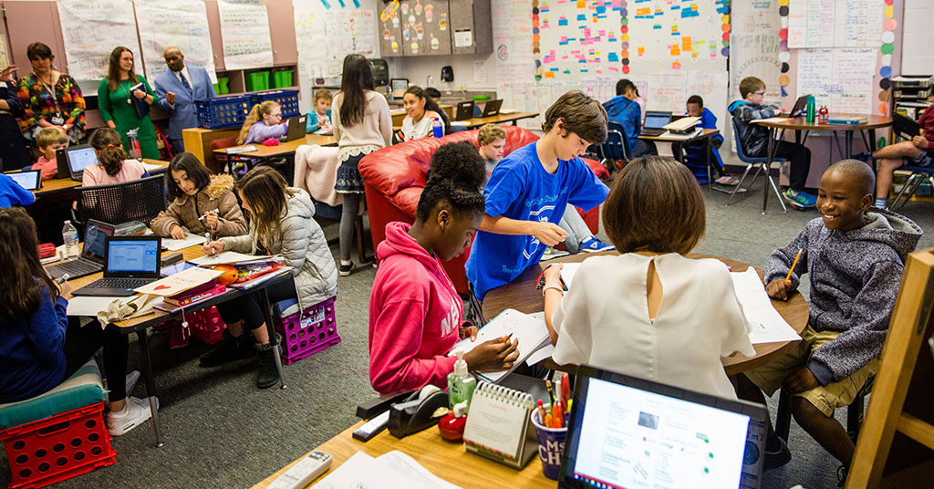 The photo is a wide shot of a fourth grade classroom. A teacher works with three students at her teacher table. In the background students are shown doing individual assignments or peer actives.