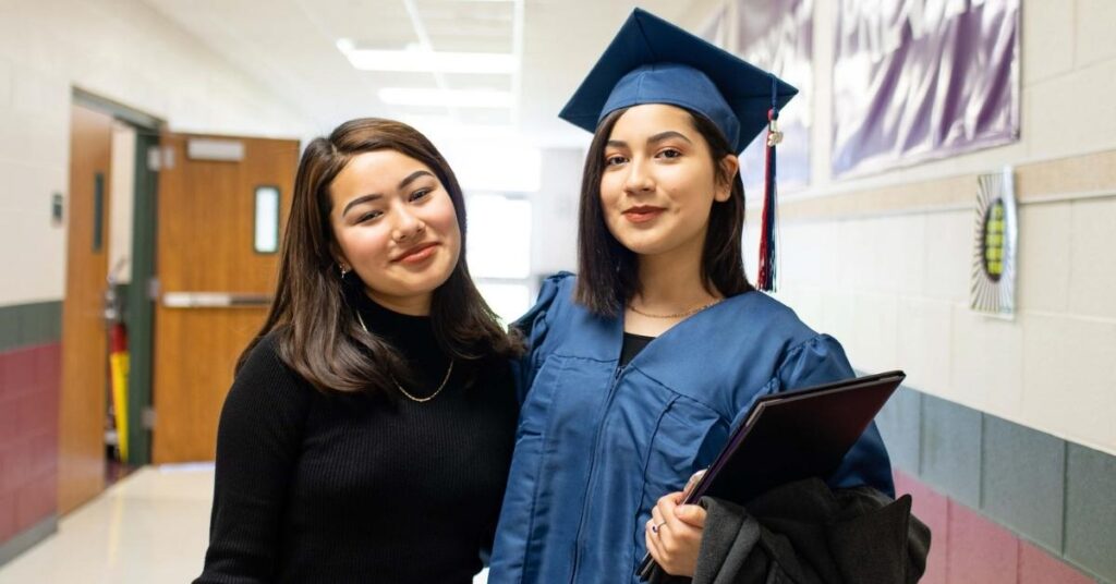 PACE graduates of two women standing side by side with their arms around each other. One is in cap and gown while the other is wearing a black long sleeve shirt.