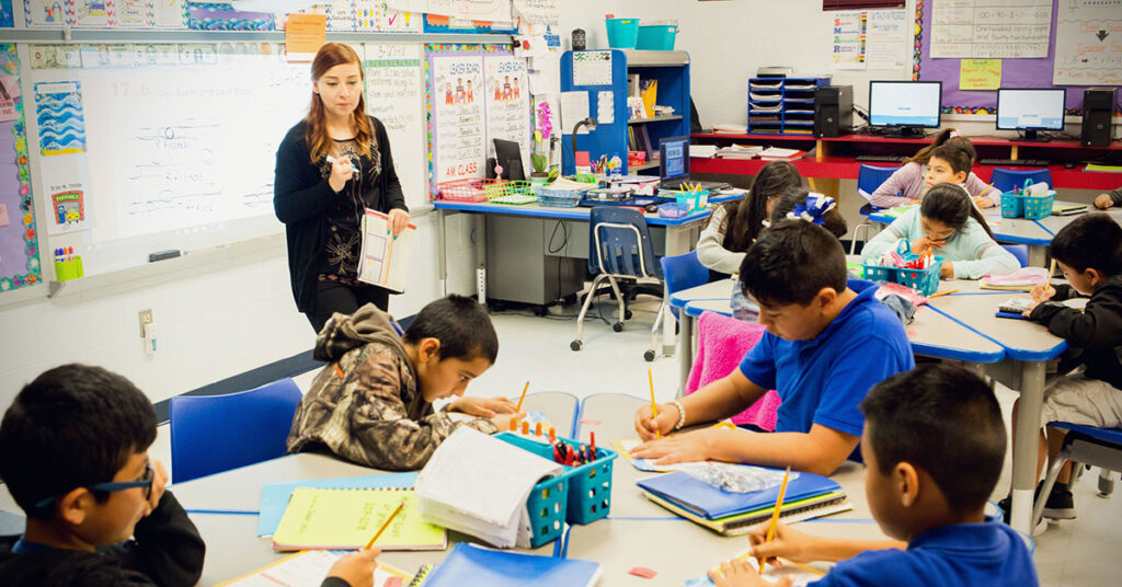 Header image of a teacher standing in front of the class teaching a diverse group of kids sitting at their desks.