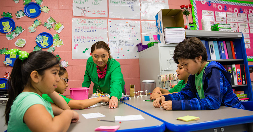 A teacher in a bright and colorful classroom leads a small group of students in a hands-on math activity.