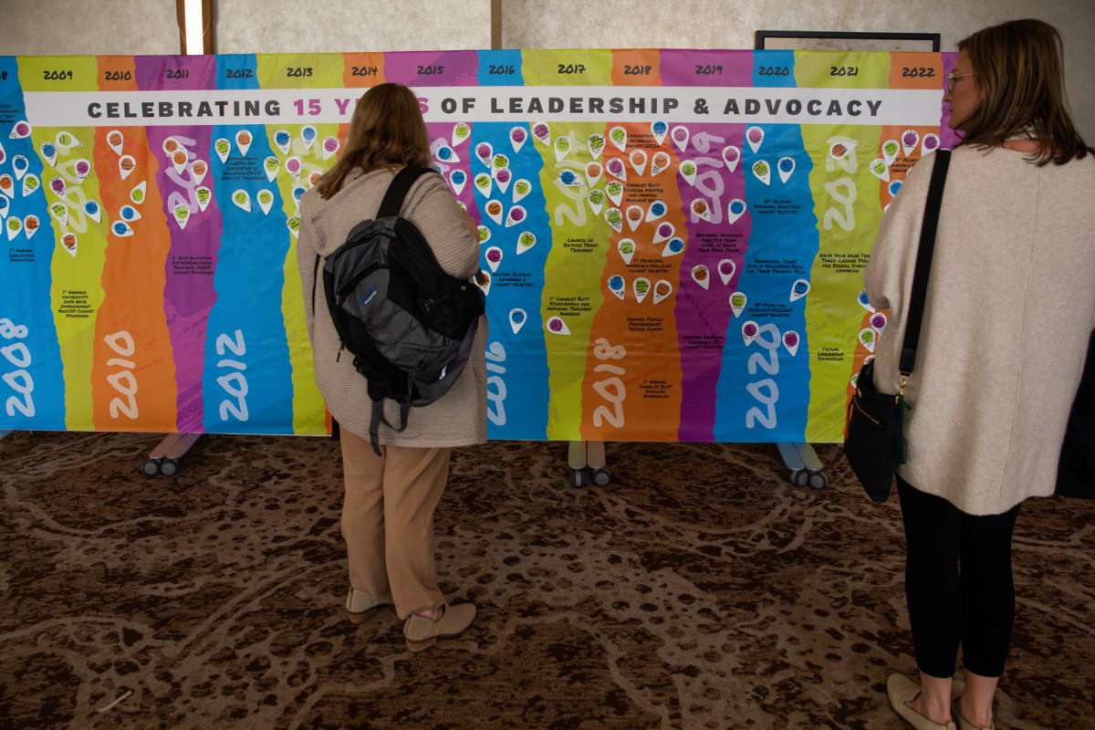 Two women stand in front of a bright colored banner titled celebrating 15 years of leadership & advocacy as they read timeline descriptions on the banner.