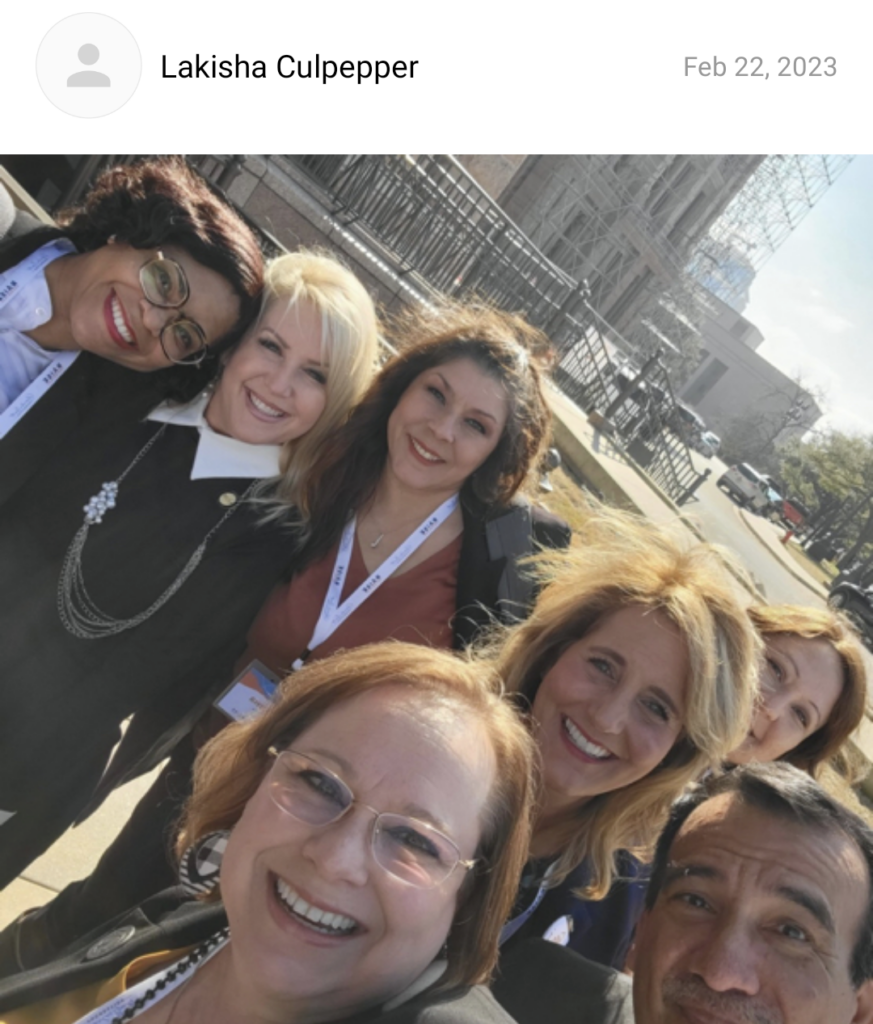 Seven educators stand close together in front of the Texas capitol smiling.