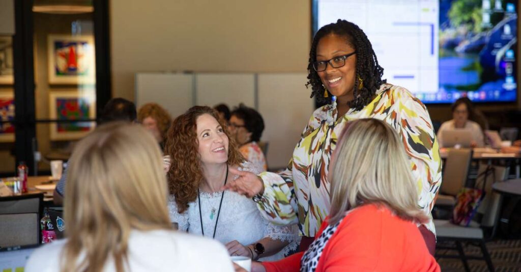 In a conference room setting, several people sit at tables. One group is up close and clear showing 3 women sitting at a table smiling and having a conversation with another woman standing and smiling. 