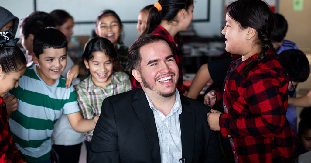 Uriel Iglesias, a 4th-grade bilingual teacher, sit in a chair as his student run up behind him for a group hug.