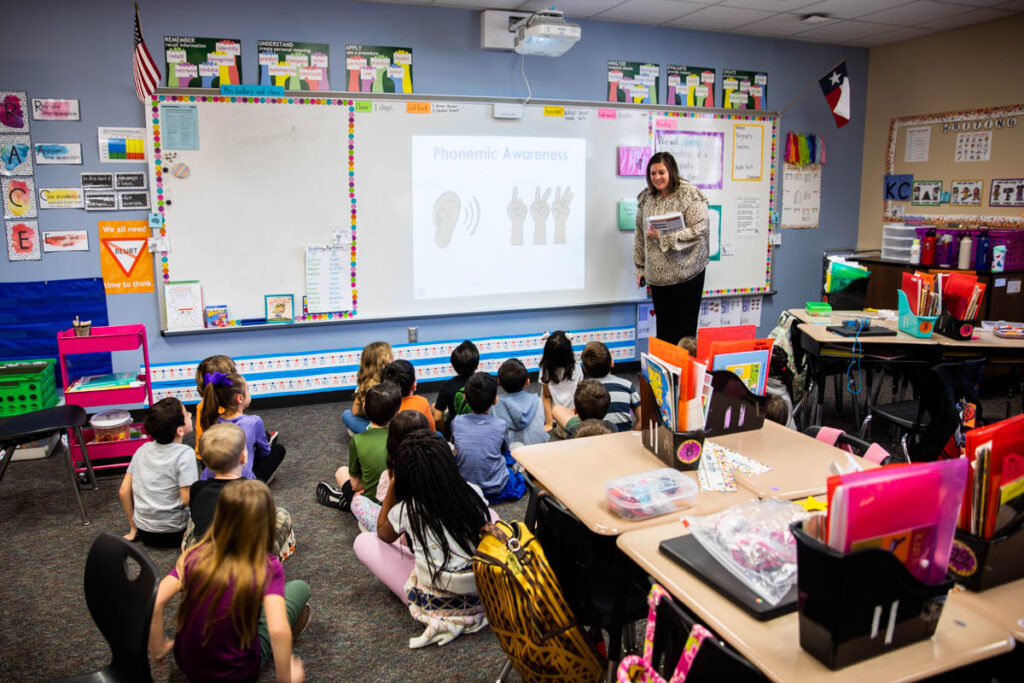 A second grade teacher guides her students in a full group lesson on phonemic awareness. In blended learning stations following the full group lesson, the students will practice these skills independently or in small groups.