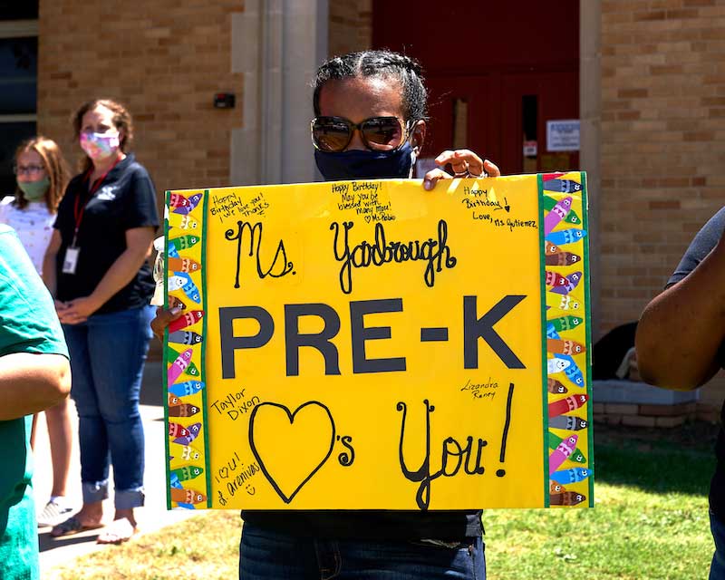 A teacher holds up a sign that reads "Ms Yarbrough Pre-K hearts you!"