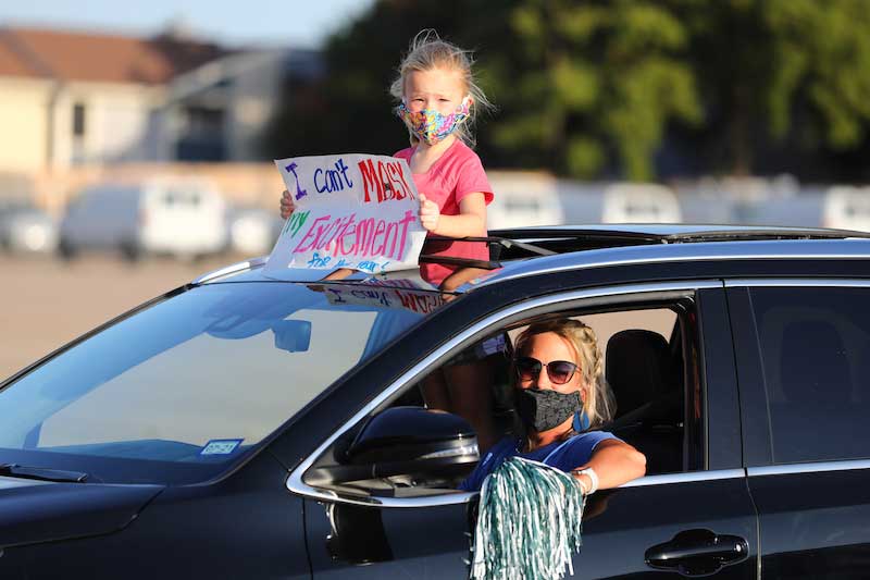 A car moves through a parade. A small child holds a sign out the car window that reads "I can't MASK my excitement."