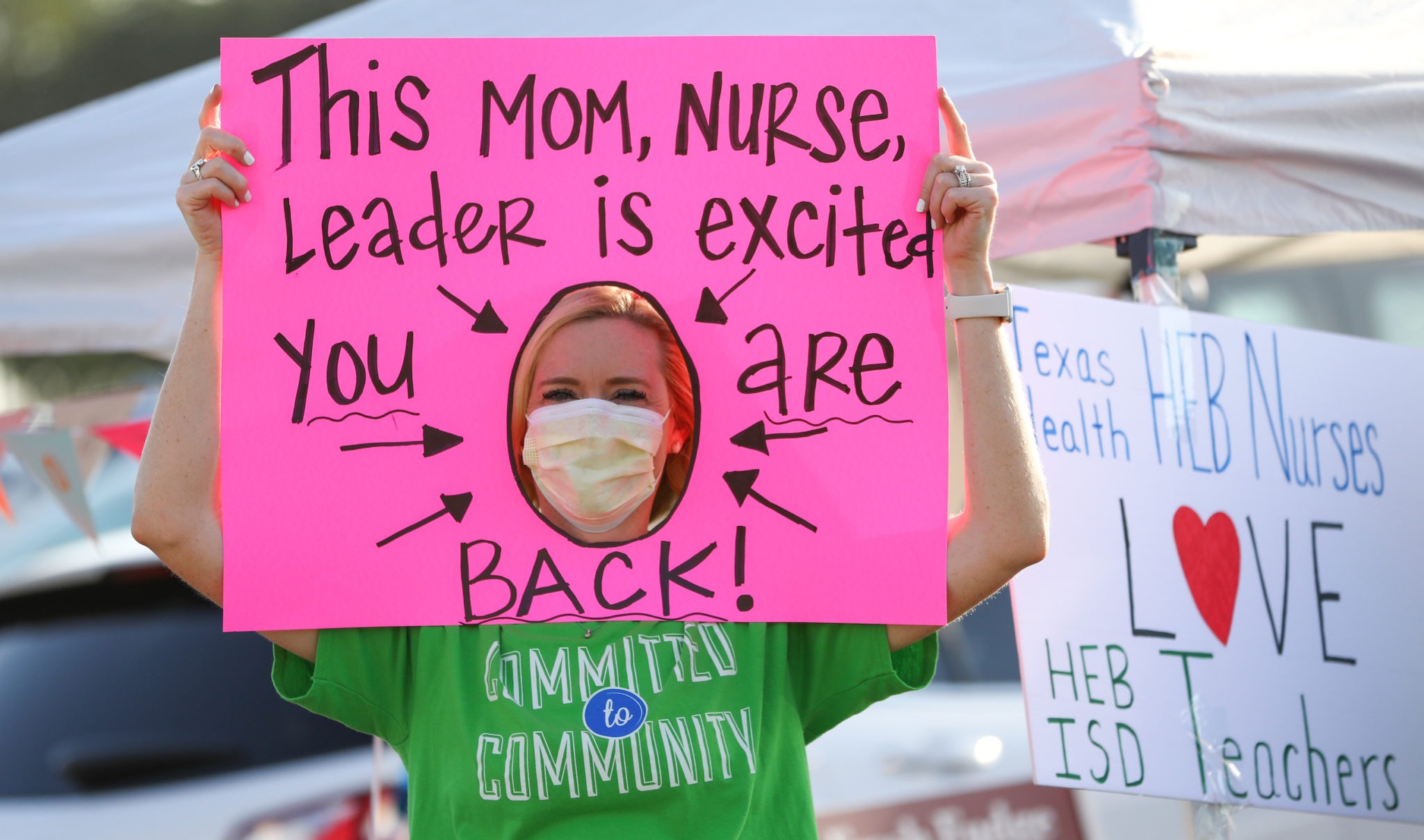 A woman in a mask holds up a pink sign that reads "This mom, nurse, leader is excited you are back!"