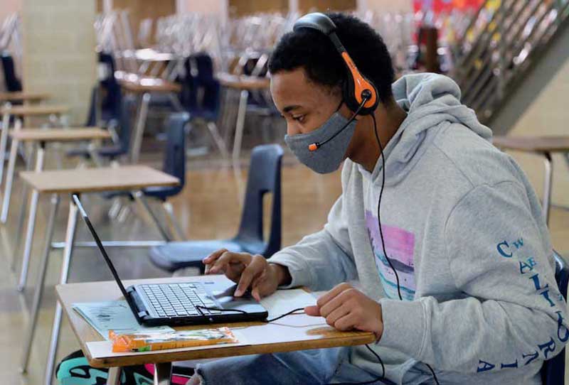 A masked high school student work at a laptop. There is a row of empty desks behind him.