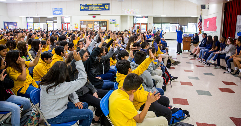 A sea of middle school students raise their hand during a Monday morning meeting in the cafeteria. The level of student engagement and school pride are high at Parades Middle School.