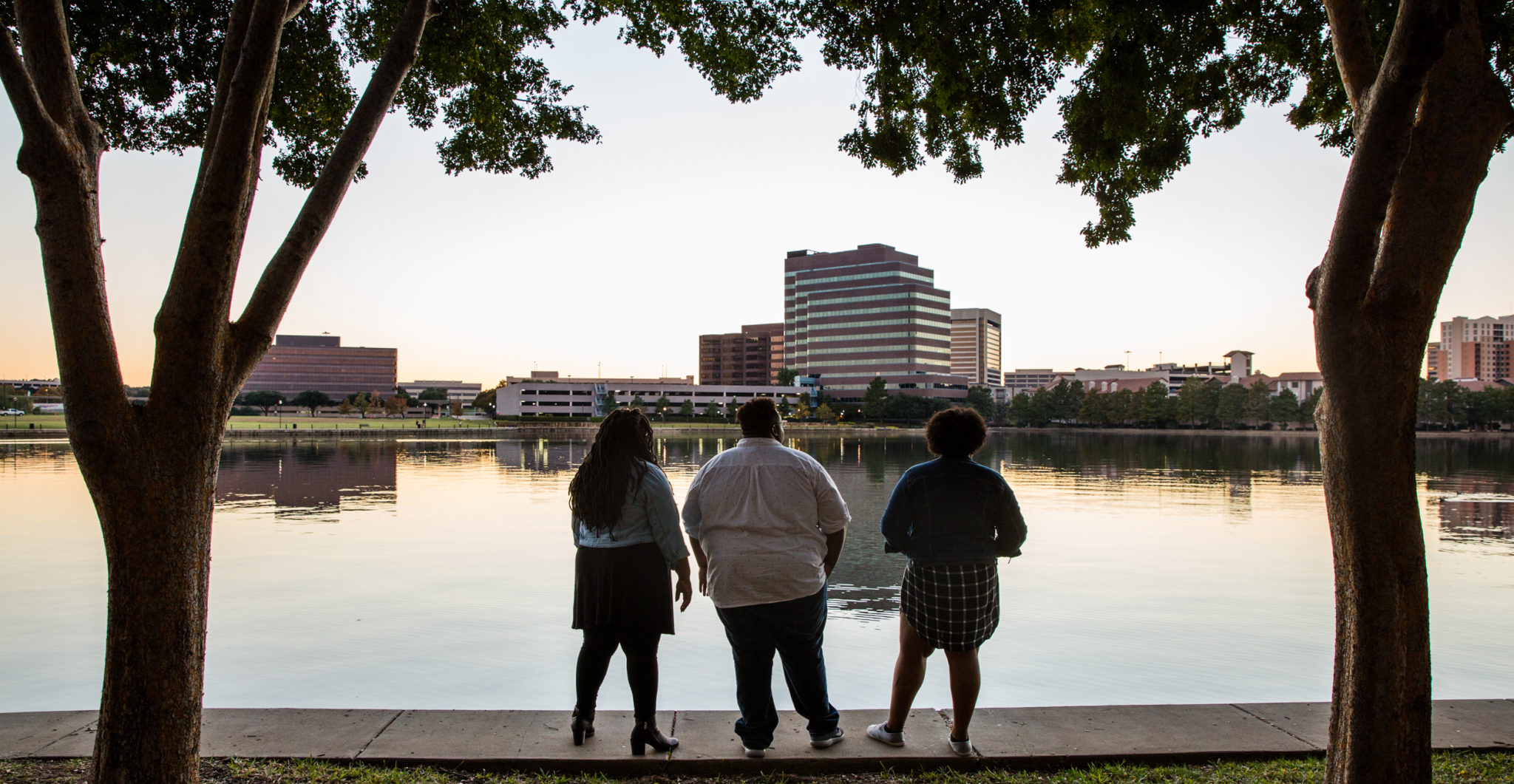 Nina, Corey, and Sherica stand in silhouette against a sunset view of the pond in front of their apartment.