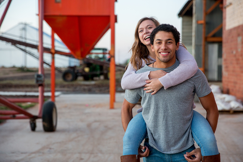 A young couple embraces into front of cotton gin in their small rural community. Both are beaming.