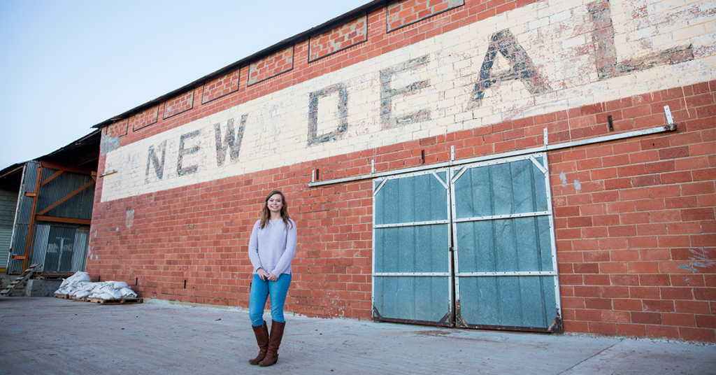 Taylor McWilliams stands in front of cotton gin in her home town with the word "New Deal Gin Co" painted on the brick exterior. 