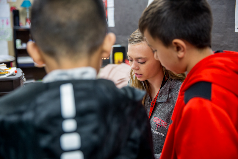 The camera peers over the shoulder of two 4th grade students who are crowded around their teacher (Taylor) to see her demonstration.