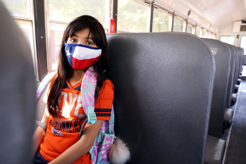 A student sits on the bus wearing a mask with the Texas flag. She looks into the camera.