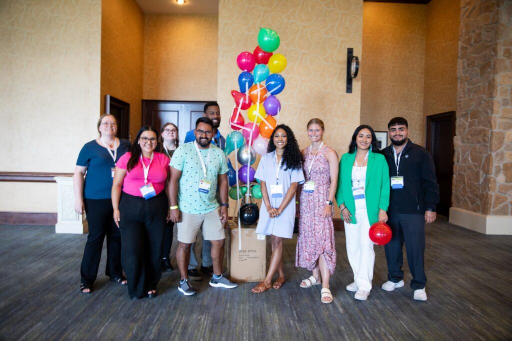 A group of 9 Charles Butt Scholar Alumni stand together smiling in front of a balloon tower they built during a team building exercise.