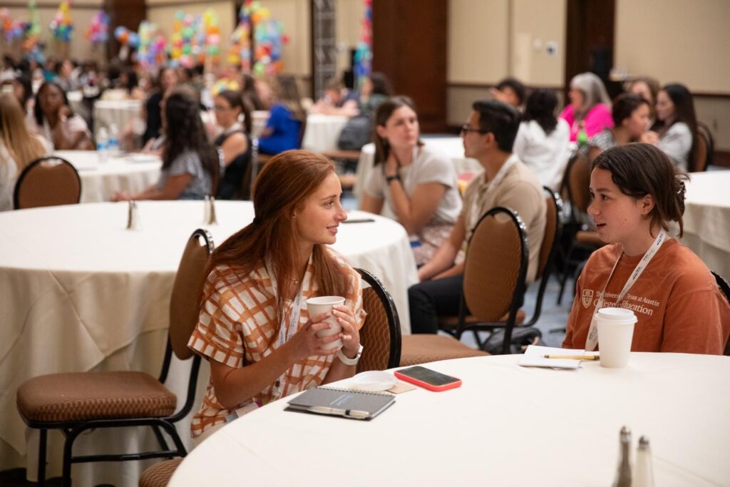 In a large conference room setting, two Charles Butt Scholar Symposium attendees sit together at a table smiling in thoughtful conversation. Behind them, out of focus, are several attendees sitting at other tables.