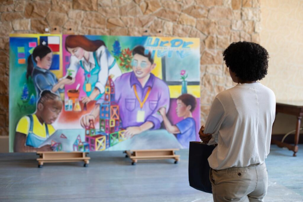 A woman stands with her back to the camera as she pensively looks at a colorful and meaningful mural of two teachers and three students working together.