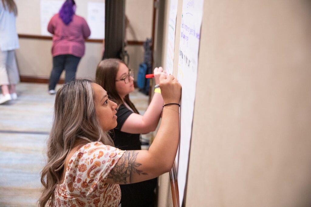 Two Charles Butt Scholar Symposium attendees stand near one another as they write on large pieces of paper with markers during a group activity.