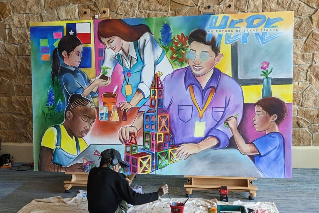 Artist, Kimie Flores, sits in front of a large mural of two teachers and three students working together. Flores adds final paint touches to the beautiful and colorful mural. The mural depicts the heart and soul of aspiring and in-service teachers.