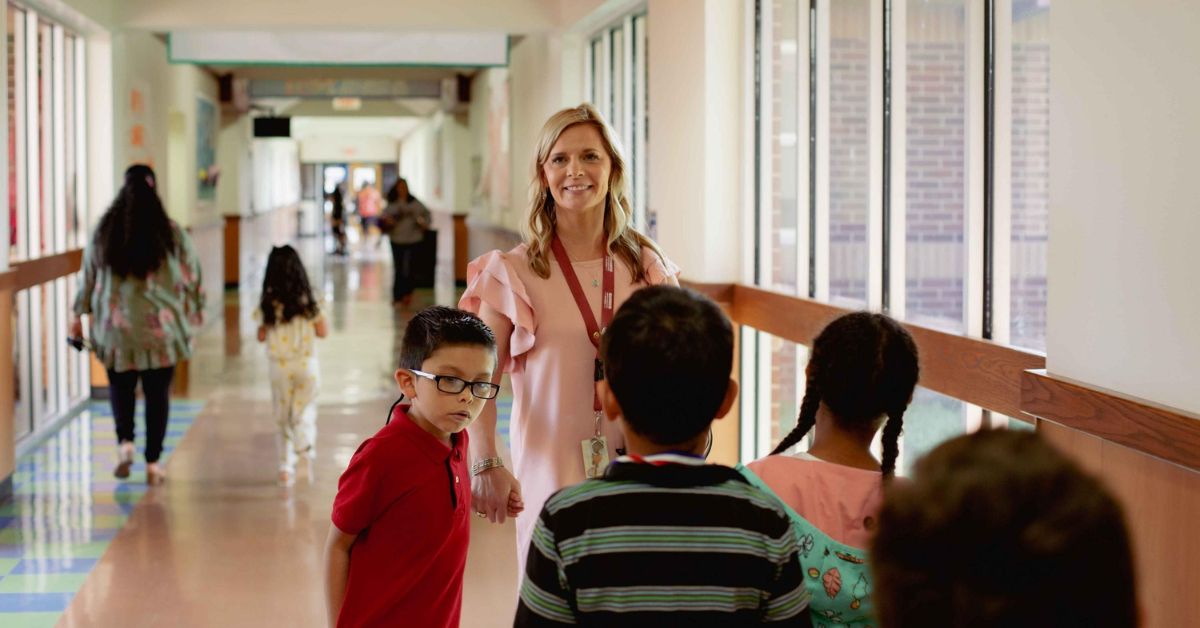 A female teacher smiles as she leads four young students through a school hallway with large sunny windows.