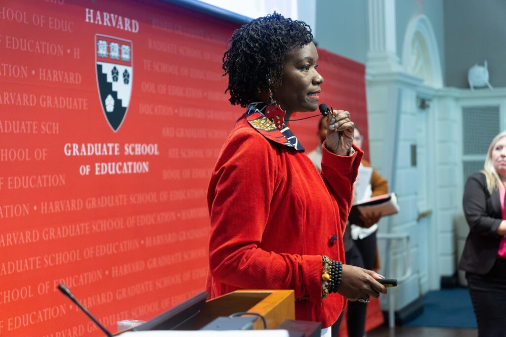 Shahara Jackson speaks into a microphone in front of a group of school leaders. There is a red backdrop behind her reading Harvard Graduate School of Education. 